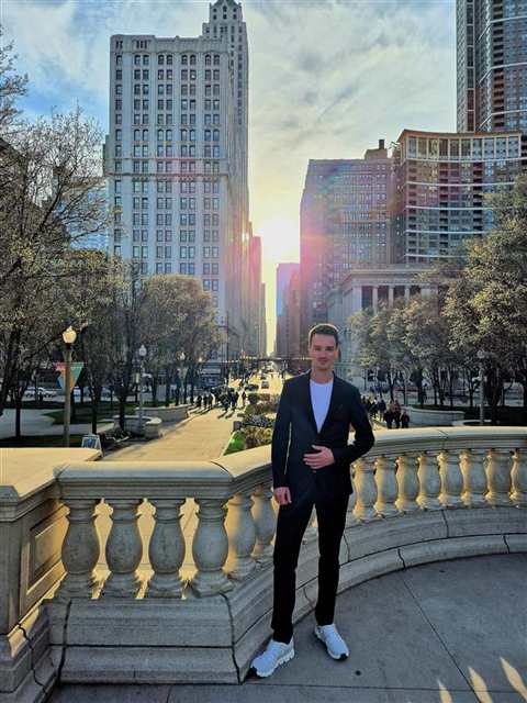 Lukas in a dark suit and white sneakers, leaning on a stone balustrade with city towers behind him at sunset